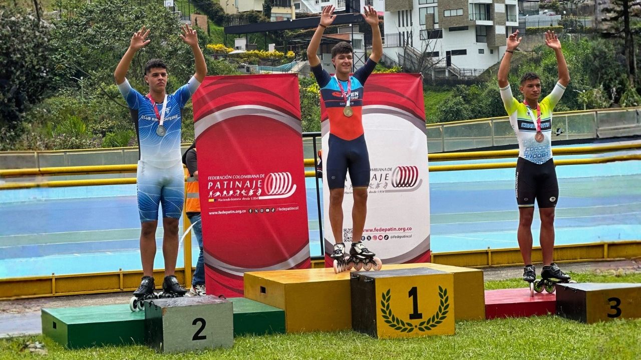 Tres patinadores de velocidad posan en un podio al aire libre durante un evento de la Federación Colombiana de Patinaje. El ganador, en el centro, viste un uniforme azul y naranja; el segundo lugar (izquierda) viste de azul claro y el tercero (derecha) de