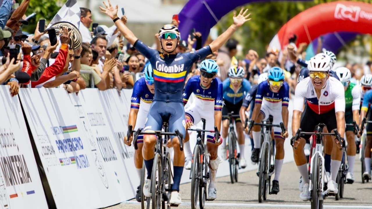 Un ciclista del equipo de Colombia cruza la línea de meta en primer lugar con los brazos en alto en señal de victoria. Viste un uniforme azul oscuro con la bandera de Colombia en el pecho. Detrás de él, otros competidores de Venezuela y Chile terminan la 