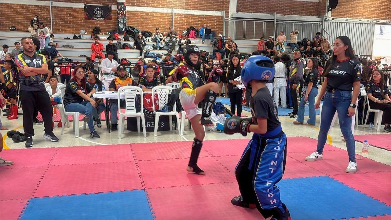 Vista de un combate de artes marciales (kickboxing o point fight) en un coliseo deportivo. Dos jóvenes competidores con equipo de protección (cascos azul y negro) se enfrentan sobre un tatami de colores rosa y azul. Una árbitra observa de cerca la acción,