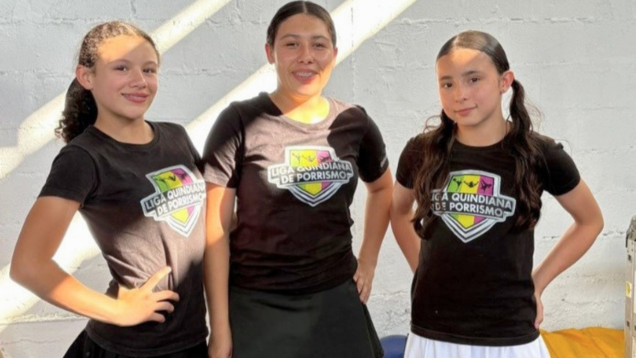 Tres jóvenes deportistas de la Liga Quindiana de Porrismo posan sonrientes frente a una pared blanca. Viste camisetas negras uniformadas con el escudo oficial de la liga en el pecho. La luz del sol entra de costado, creando un ambiente cálido y profesiona