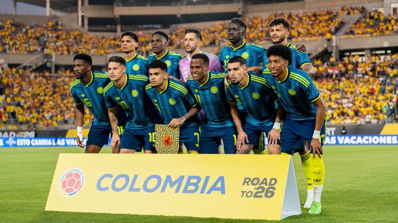 Fotografía oficial de la alineación titular de la Selección Colombia masculina de fútbol en un estadio lleno. Los jugadores visten el uniforme alternativo de color azul oscuro con detalles en amarillo neón. En primer plano, una valla publicitaria amarilla