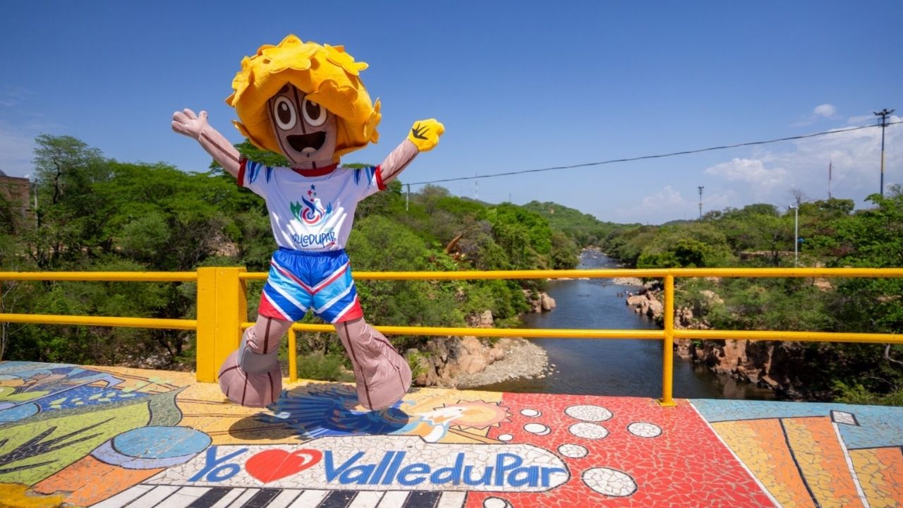 Tobalito, la mascota oficial de los Juegos Bolivarianos Valledupar 2022, un personaje con cabello amarillo en forma de rayos de sol, saltando con los brazos abiertos sobre un puente decorado con mosaicos coloridos. Al fondo se aprecia el río Guatapurí rod