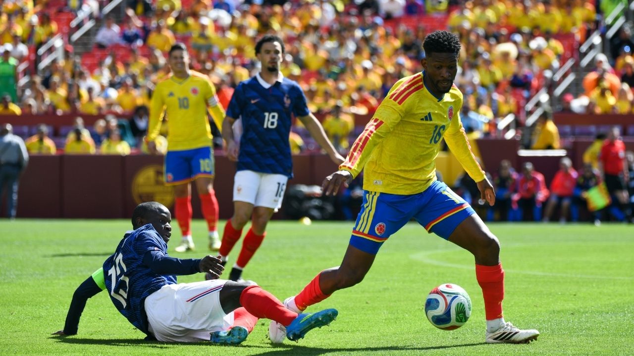 Un jugador de la selección de Colombia con el dorsal 16 controla el balón durante un partido de fútbol, ​​mientras un jugador de Francia (dorsal 23) intenta quitarle la pelota barriéndose en el césped. Al fondo se ve a James Rodríguez y otros jugadores en