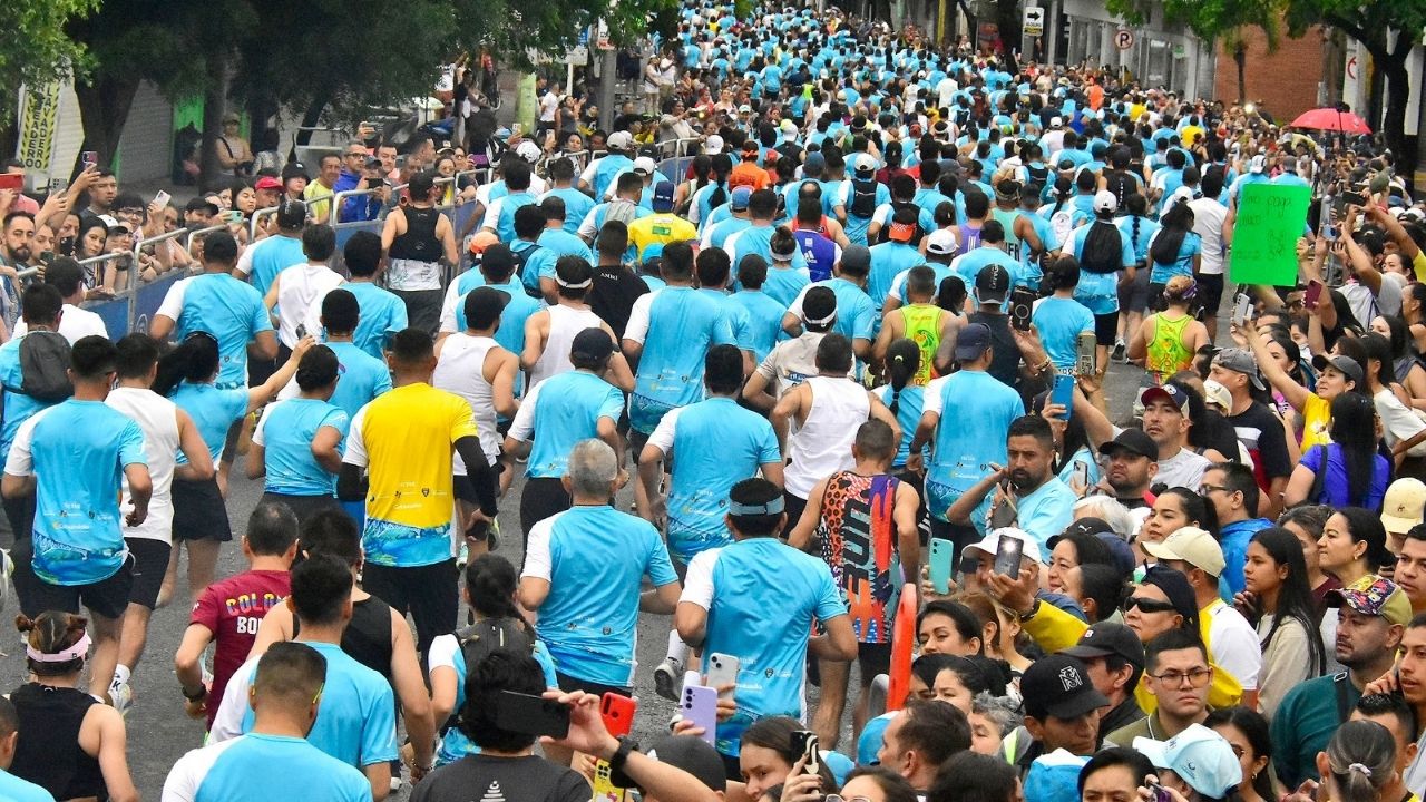 Una toma aérea desde atrás de una gran multitud de corredores participando en un maratón. La mayoría de los participantes visten camisetas técnicas de color azul claro, creando un "mar" de gente que llena una avenida de la ciudad. A los lados, se observan