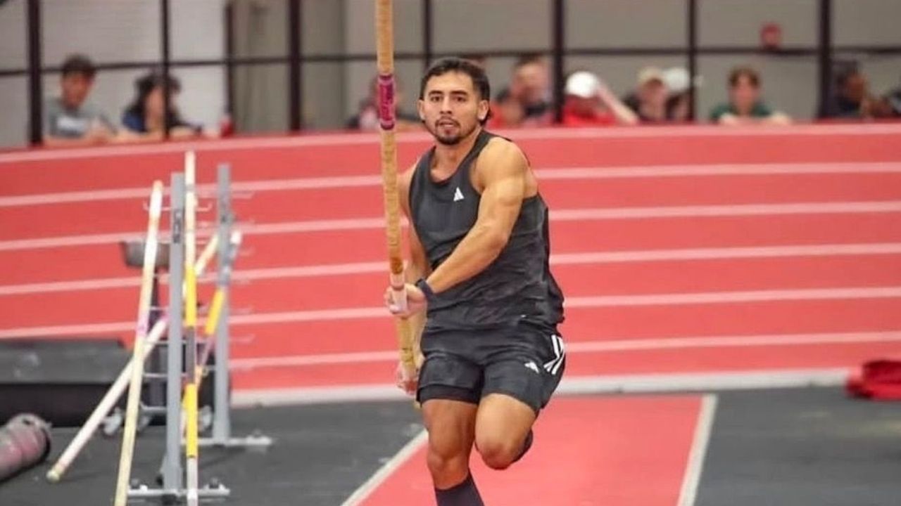 Un atleta masculino de salto con garrocha en plena carrera de aproximación sobre una pista roja de atletismo bajo techo. El deportista viste uniforme negro, sostiene la garrocha con ambas manos y muestra una expresión de total concentración. Al fondo se o