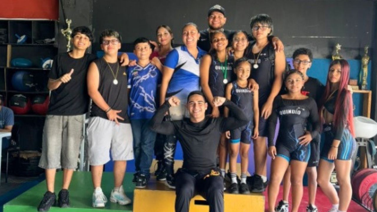 Una fotografía grupal de un equipo deportivo de levantamiento de pesas en un gimnasio. El grupo, compuesto por jóvenes y adultos, posa sonriente sobre un podio de tres niveles. La mayoría viste ropa deportiva de color negro; una mujer en el centro lleva u
