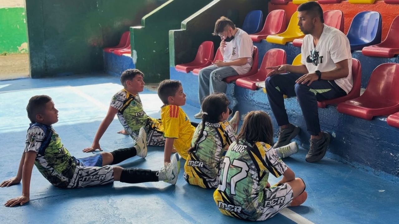 Una toma de ángulo bajo que captura a un entrenador adulto dando instrucciones a un grupo de cinco niños atletas sentados en una cancha deportiva azul de interior. El entrenador, que lleva una camiseta blanca y pantalones negros, está sentado en las grada