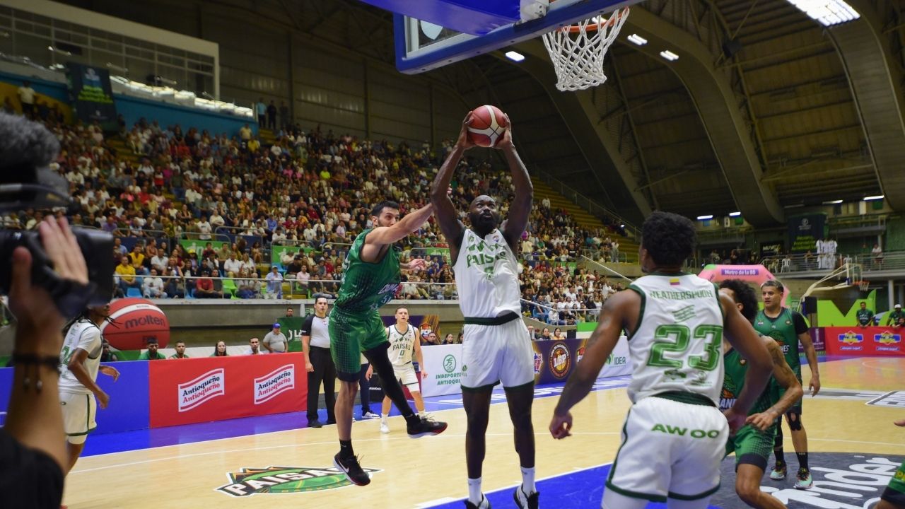 Un jugador de Paisas captura un rebote cerca del aro en medio de la defensa rival. La toma amplia muestra la cancha de baloncesto, el cuerpo técnico y las tribunas llenas de espectadores en un ambiente de competencia profesional.