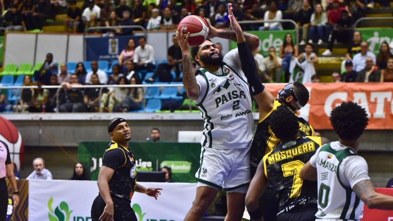 Jugador del equipo Paisas (blanco) salta para lanzar al aro mientras es bloqueado por un defensor. Alrededor, otros jugadores observan la jugada en un coliseo con público al fondo durante un partido de la liga profesional.