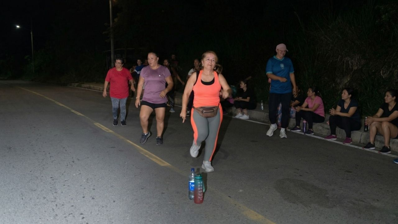 Grupo de mujeres practicando atletismo en una vía pública durante la noche. Una mujer en primer plano viste camiseta naranja y leggings grises con franja naranja, mientras otras compañeras observan o corren detrás de ella.