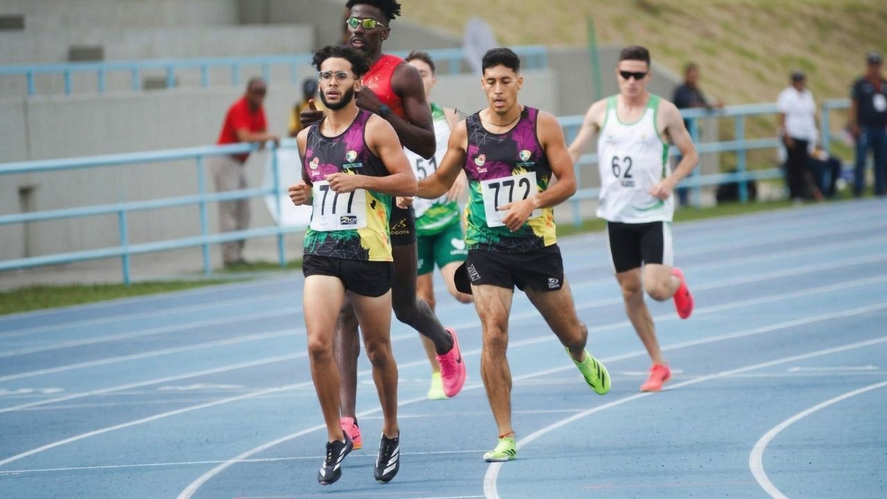 Varios corredores en una pista de atletismo azul durante una competencia. En primer plano, dos atletas con uniformes oscuros y coloridos, dorsales 777 y 772, concentrados en la carrera bajo la luz del día.