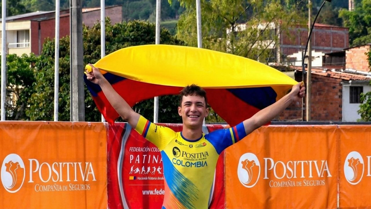 Patinador juvenil con el uniforme de la Selección Colombia "Positiva" celebrando con los brazos en alto y la bandera nacional tras sus hombros. Al fondo, vallas publicitarias y vegetación urbana.