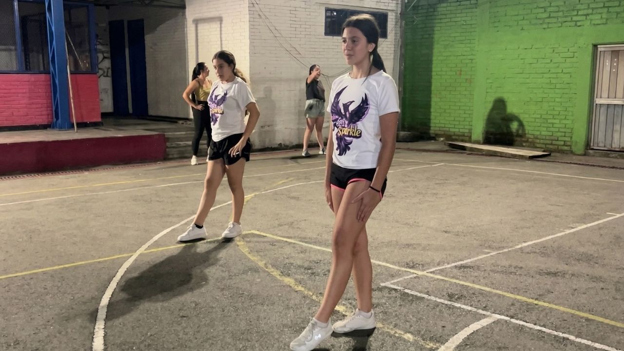 Dos jóvenes con camisetas blancas de "Fénix Sparkle" practicando una rutina deportiva nocturna en una cancha múltiple. Al fondo se ven otras compañeras y paredes de ladrillo iluminadas.