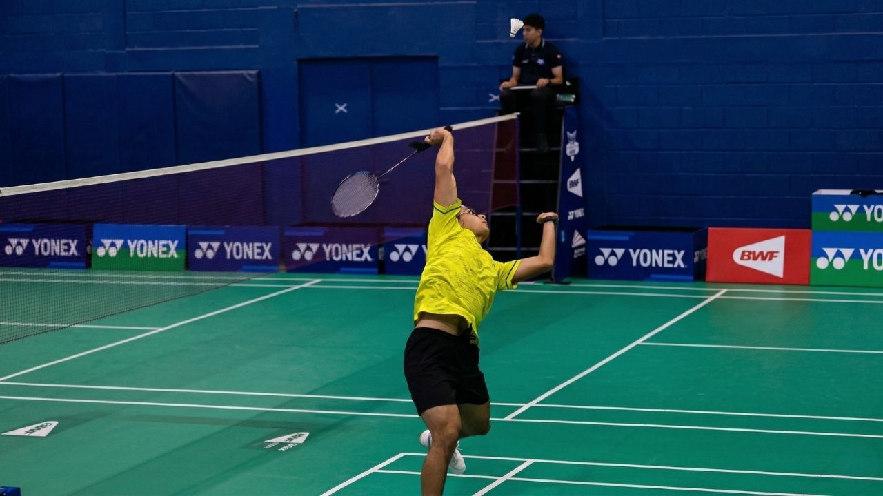Jugador de bádminton con uniforme amarillo neón en pleno salto realizando un remate potente. Se aprecia la cancha verde, la red y un juez de silla al fondo sobre una pared azul.