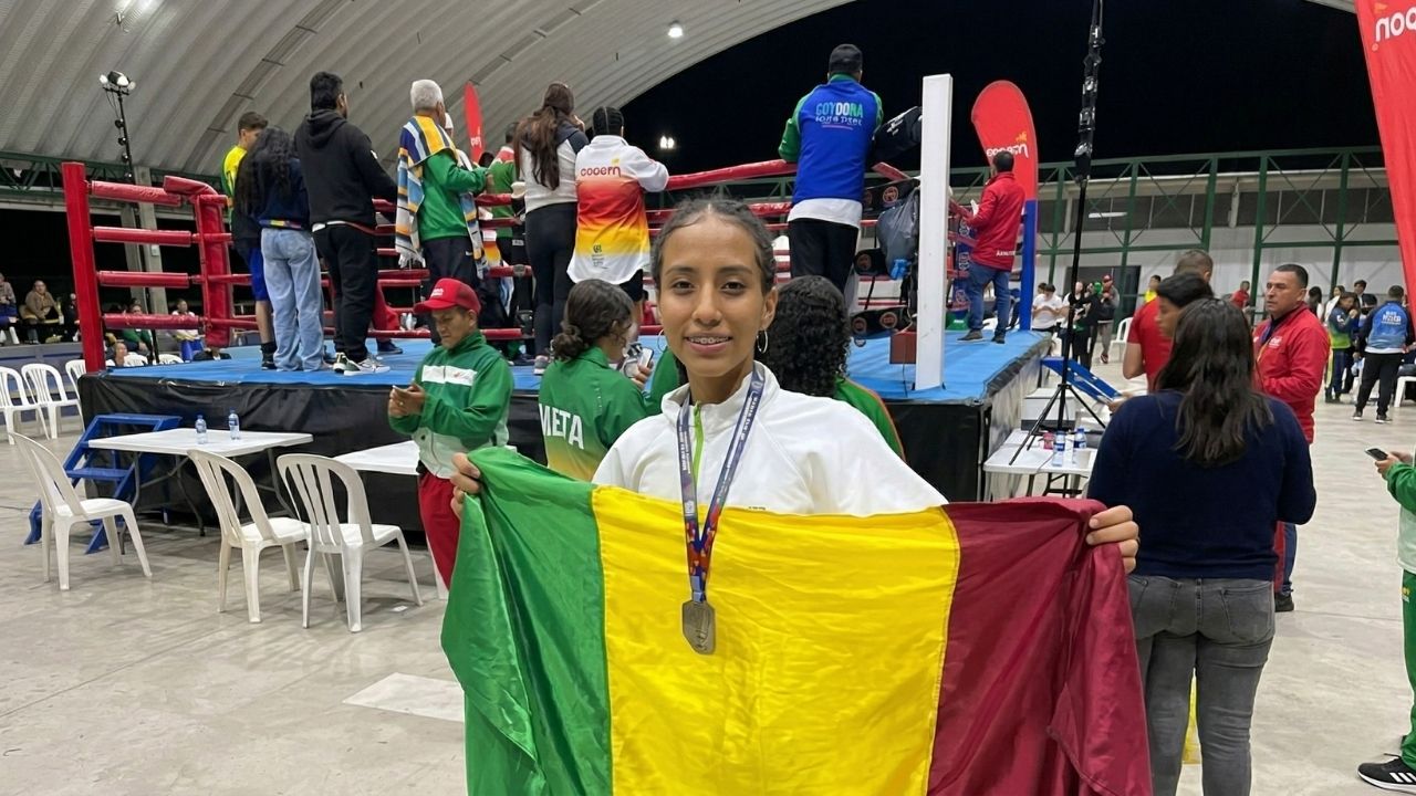 Joven deportista sonriente con medalla al cuello, sosteniendo la bandera de Colombia. De fondo, un ring de boxeo en un coliseo con público y deportistas con uniformes de ligas regionales.