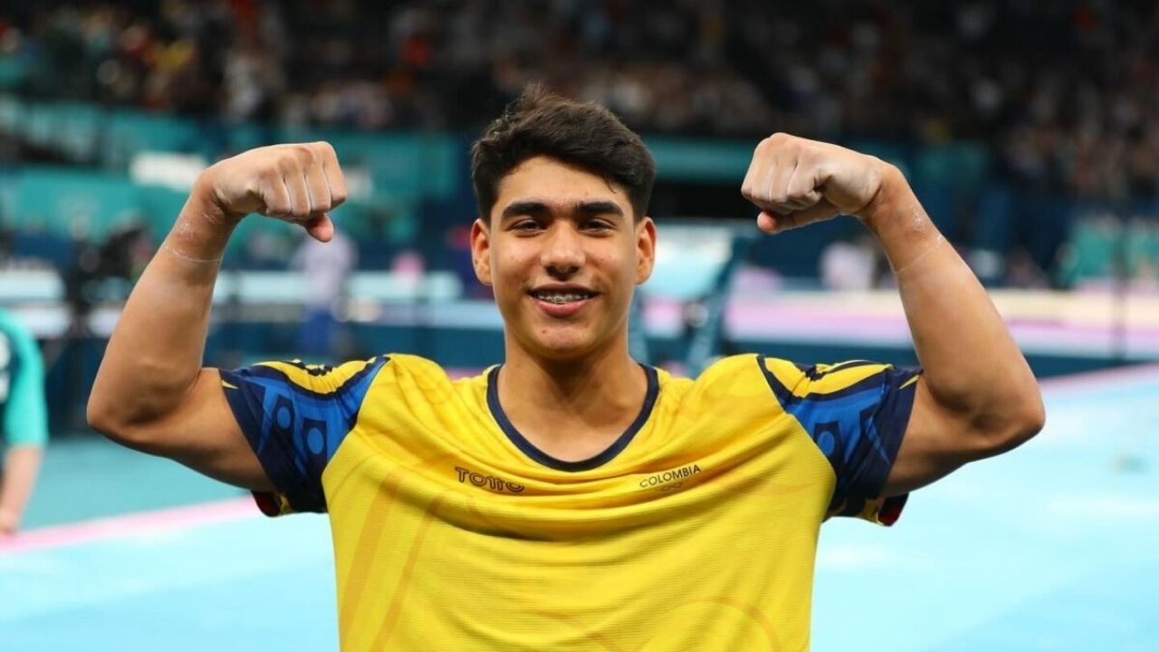 Primer plano del gimnasta Ángel Barajas con el uniforme amarillo de Colombia, celebrando con los brazos flexionados mostrando fuerza y una sonrisa. De fondo, un gimnasio de competición desenfocado. Imagen de alta calidad y estilo editorial.