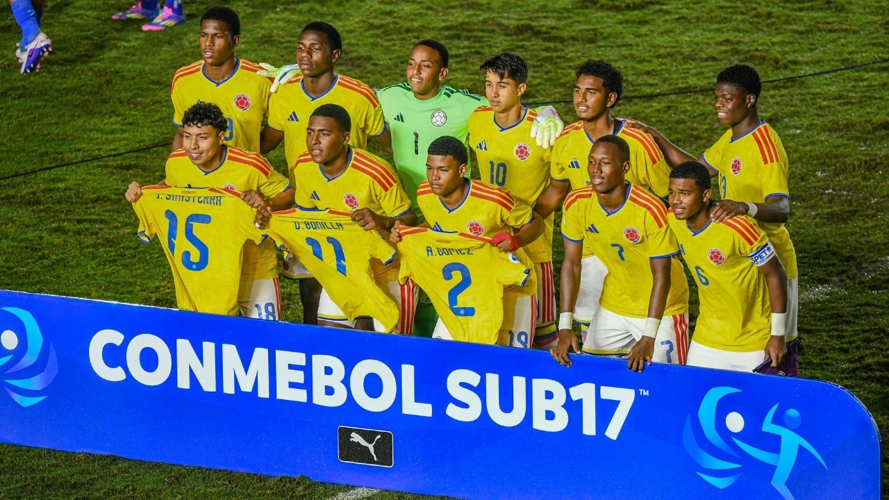 La selección de fútbol Sub-17 de Colombia posa en grupo tras una valla de CONMEBOL. Tres jugadores sostienen camisetas con los nombres de compañeros ausentes (J. Santana, D. Bonilla y A. Gómez) en un gesto de apoyo.