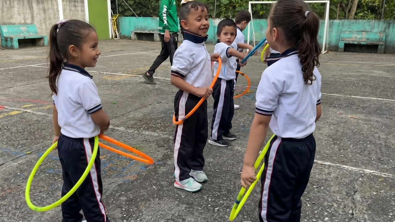Cuatro niños con uniformes escolares (polos blancos, pantalones oscuros con franja) juegan con aros en un patio de cemento. La niña del centro sonríe con un aro naranja. Detrás, hay otros niños, un adulto de verde y bancas.