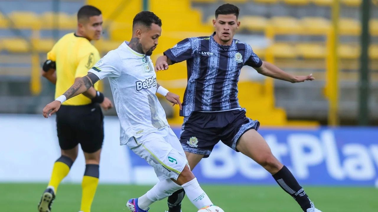 Jugador de blanco (BetPlay) driblea el balón ante la presión de un rival de uniforme azul oscuro en un estadio. Acción de fútbol profesional masculino en campo abierto con árbitro al fondo.