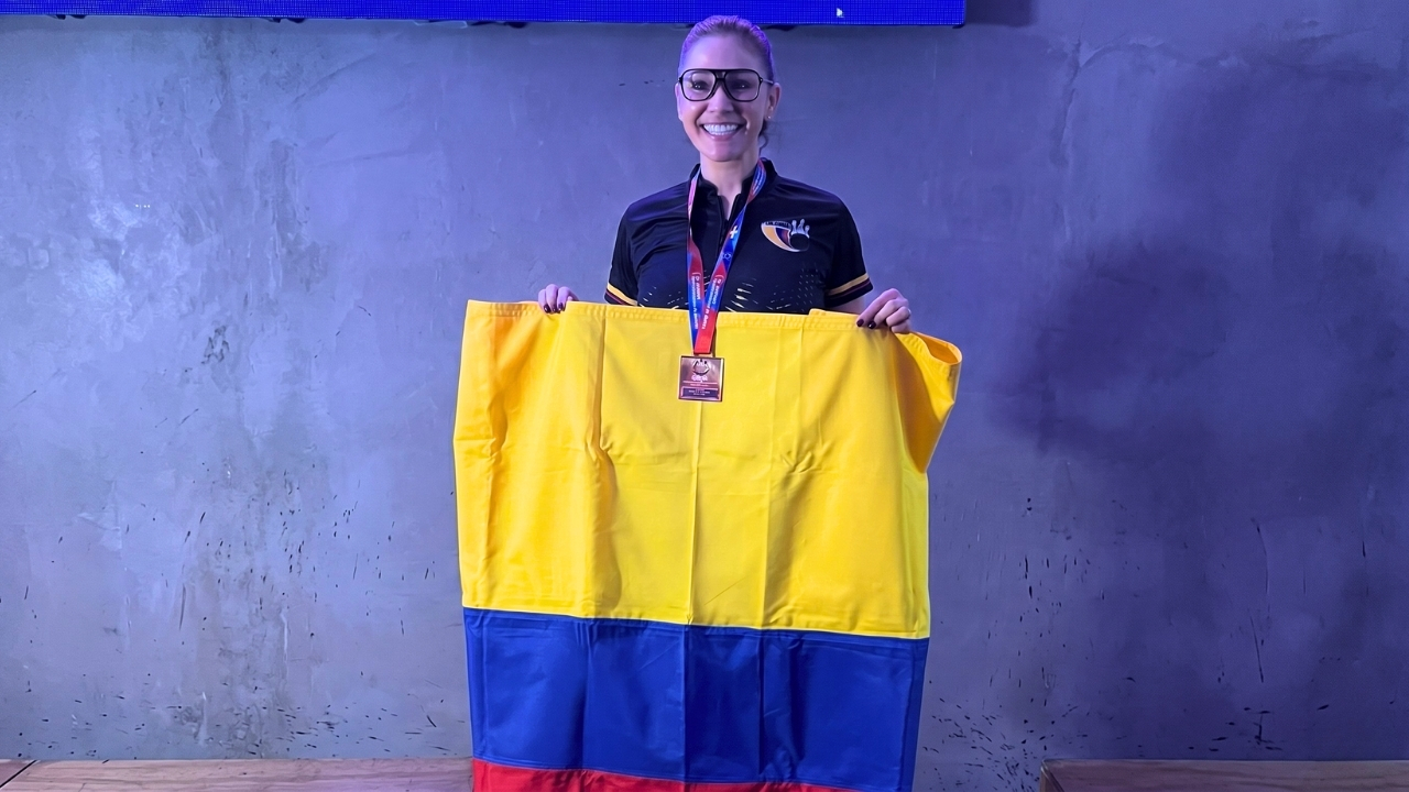 Una mujer sonriente con gafas deportivas y una medalla al cuello sostiene con orgullo una bandera de Colombia (amarillo, azul y rojo) frente a una pared de textura rústica y fondo grisáceo.