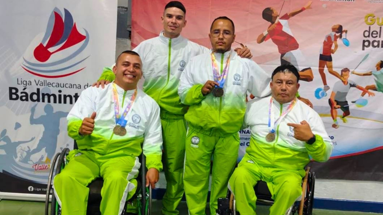 Cuatro deportistas de bádminton adaptado posan con sus medallas. Dos están en sillas de ruedas y dos de pie, todos vistiendo uniformes blancos y verdes de la delegación del Quindío frente a un banner del evento.