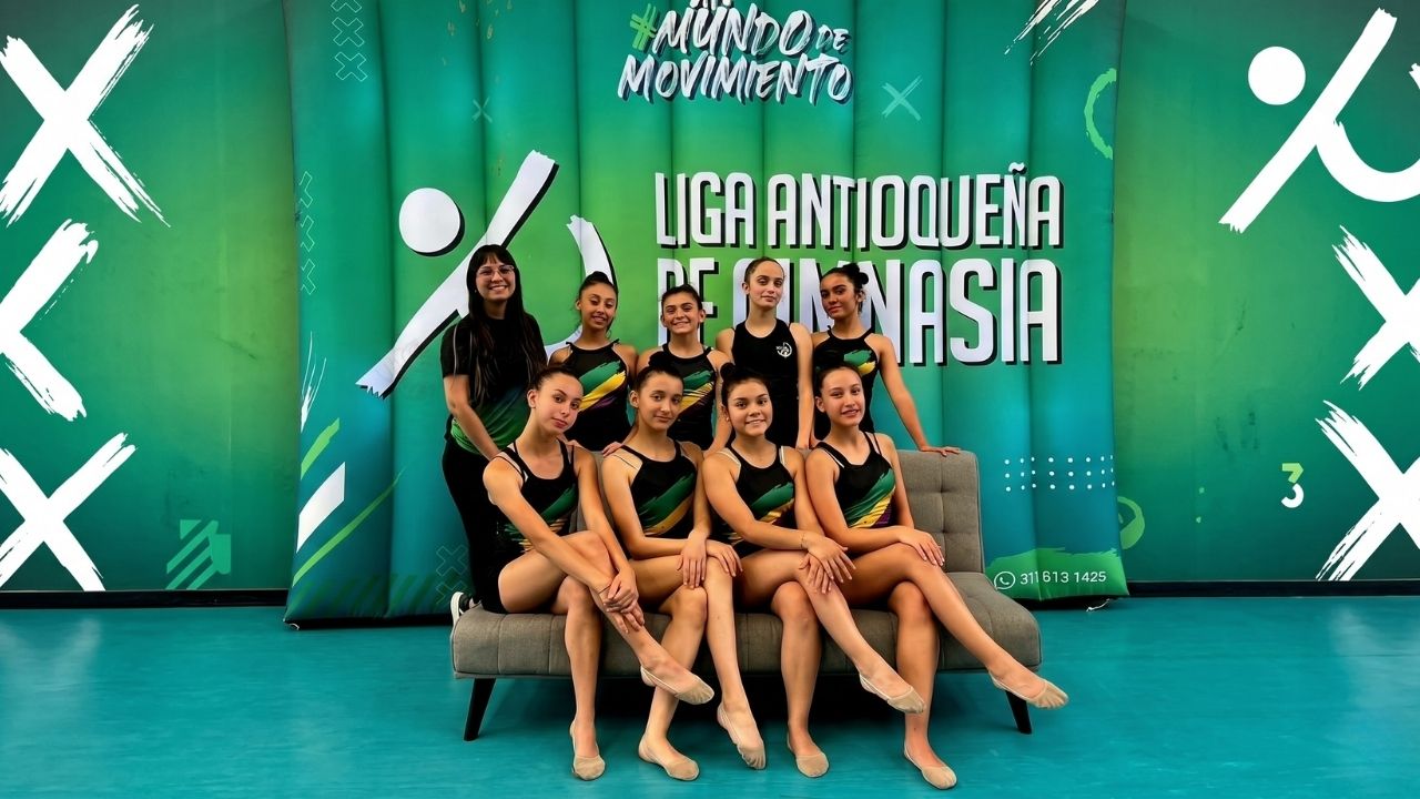 Nueve integrantes de la Liga Antioqueña de Gimnasia posan sonrientes en un sofá gris frente a un fondo verde con el lema "#MundoDeMovimiento". Visten uniformes deportivos negros con detalles verdes y amarillos en un ambiente de entrenamiento.